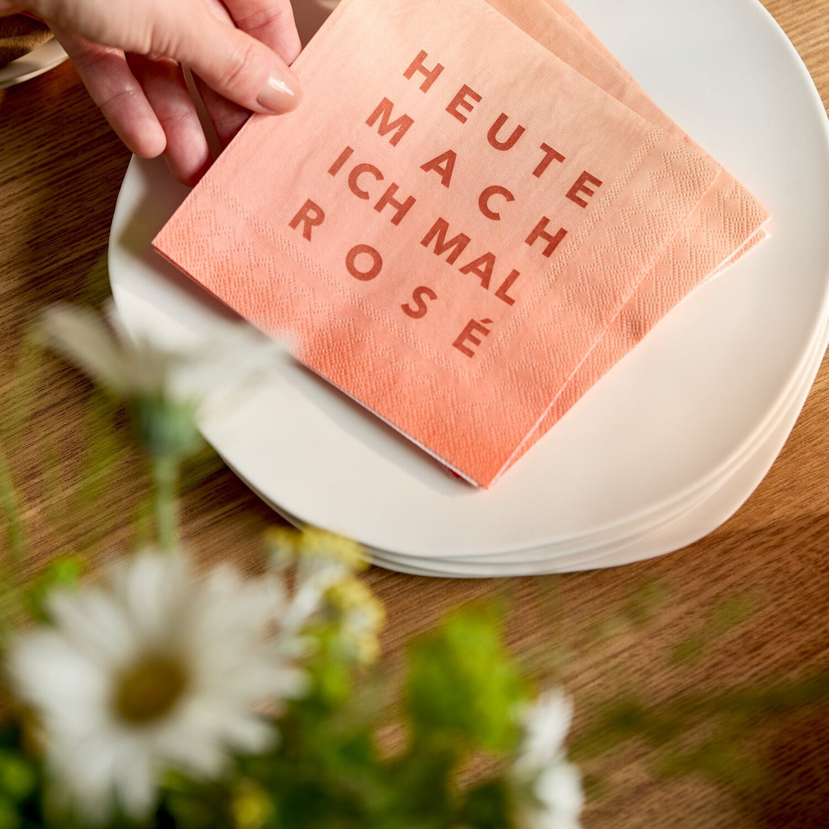 Orange Cocktailserviette 25x25 cm, gefaltet auf Teller, Hand hält Serviette mit Rosé-Text