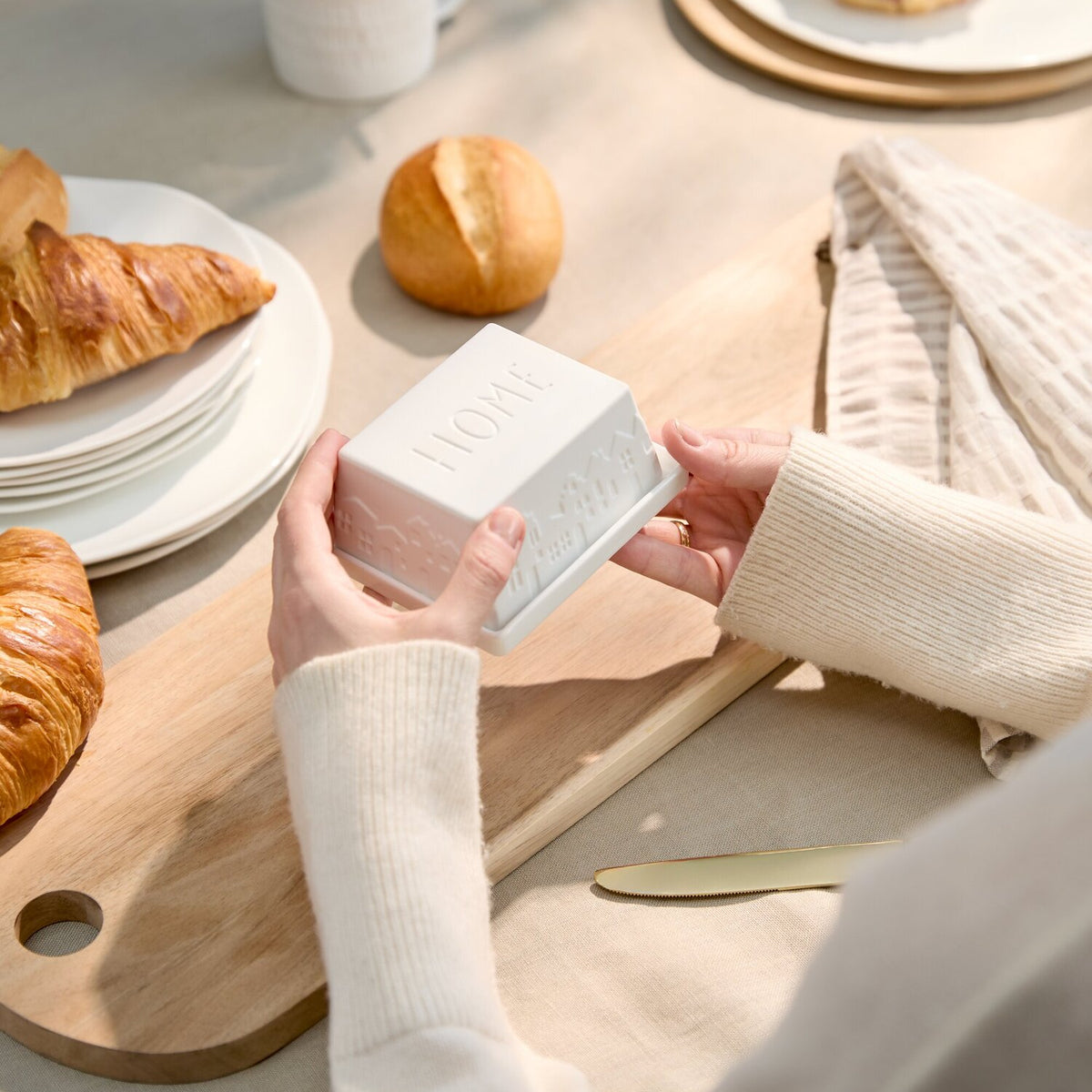 Person hält weiße Butterdose „SWEET HOME“ auf Holzbrett neben Tasse