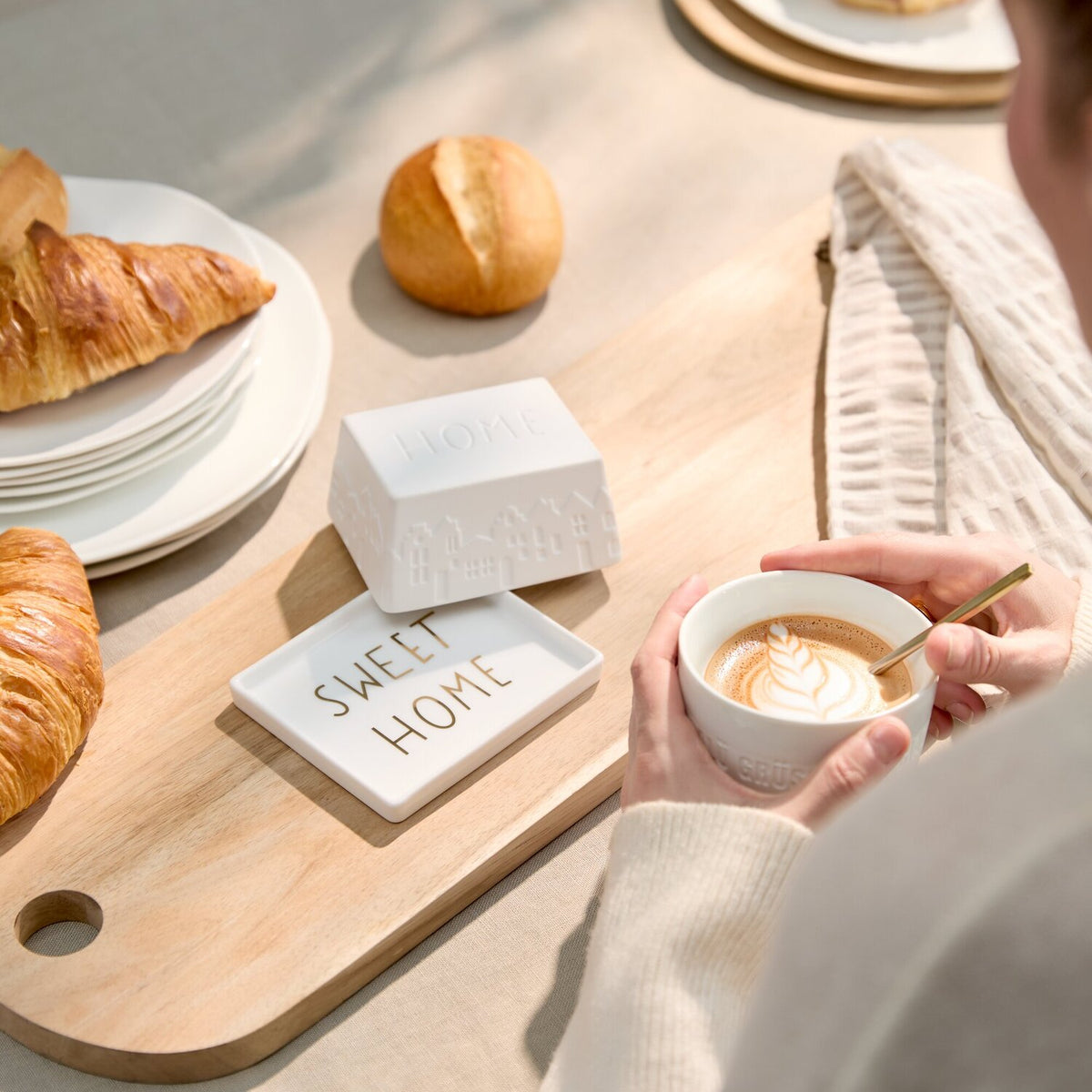 Weiße Butterdose „SWEET HOME“ auf Holzbrett, Person hält Tasse mit Milchschaum
