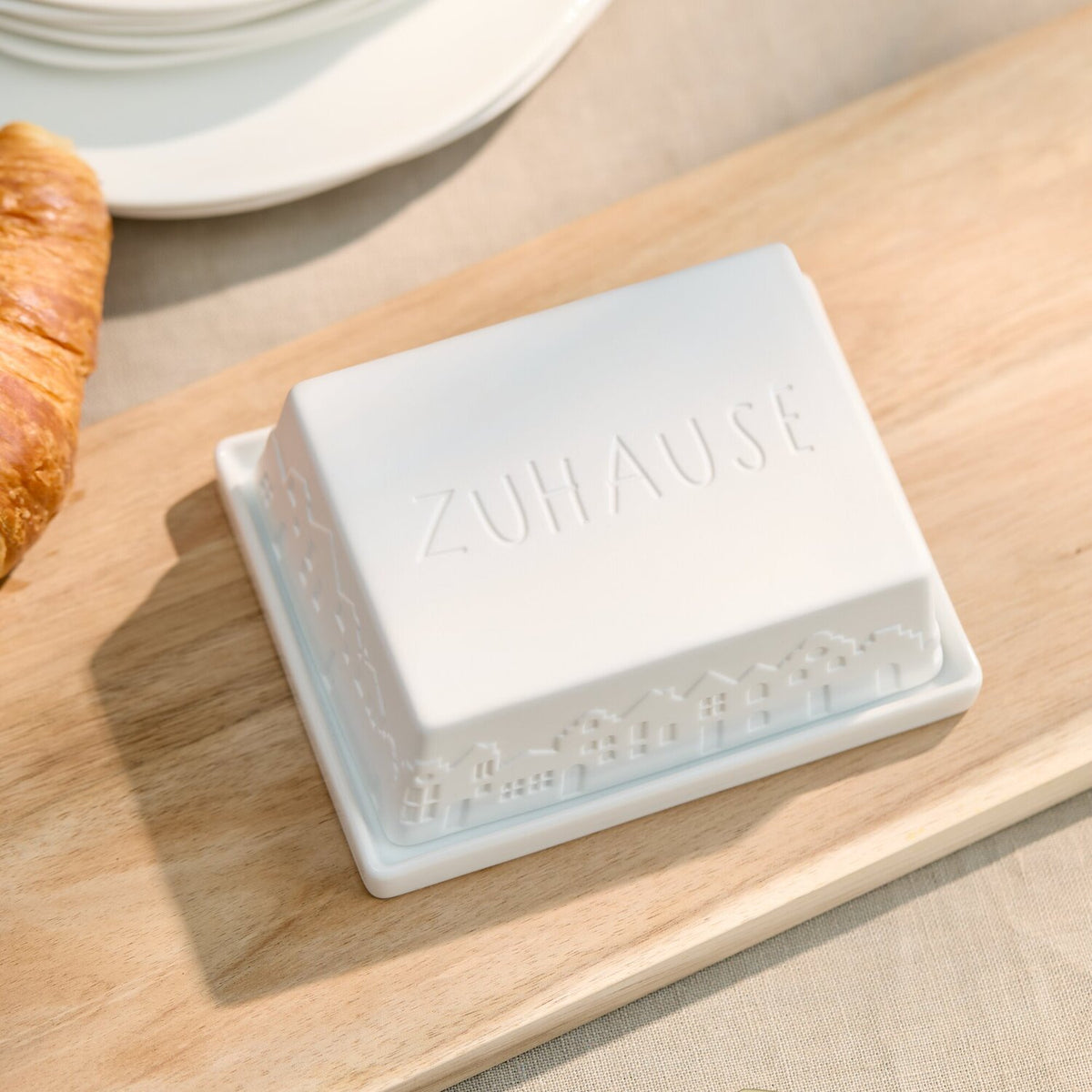 Weiße Butterdose auf Holzbrett mit geprägtem „ZUHAUSE“ auf dem Deckel