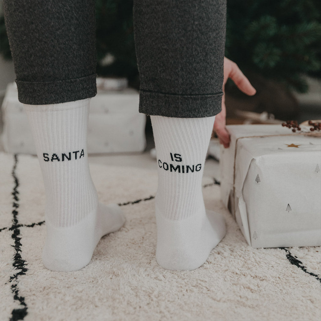 Weiße Socken mit Aufdruck „SANTA“ und „IS COMING“ am Schaft.