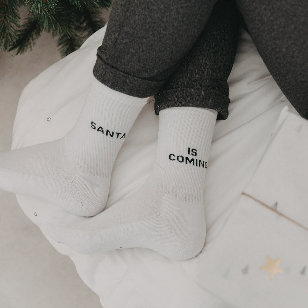 Weiße Socken am Fuß mit Aufdruck „SANTA“ und „IS COMING“.