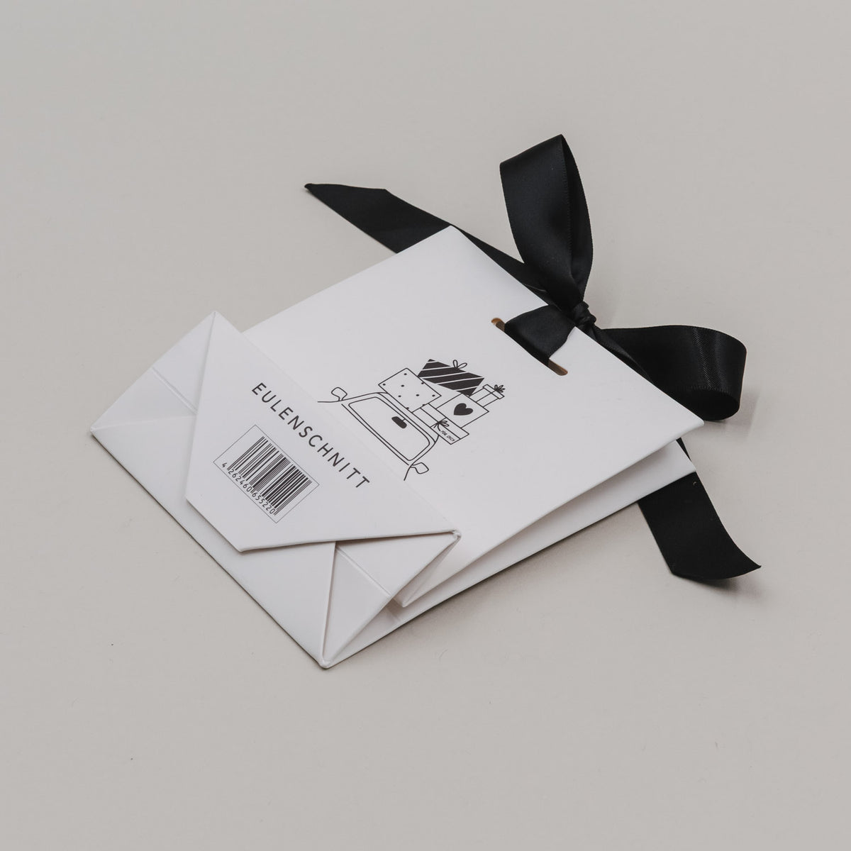 Weiße Geschenktüte mit schwarzem Band; Rückseite mit Text „EULEN SCHNITT“ und Barcode