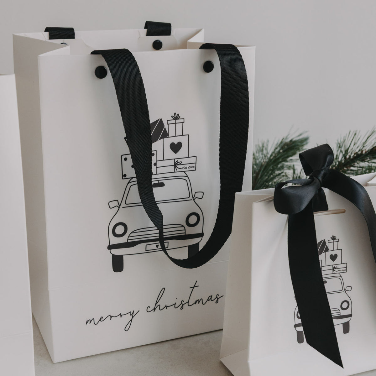 Zwei weiße Geschenktüten nebeneinander, schwarzes Trageband und Weihnachtsauto-Motiv im Vordergrund.
