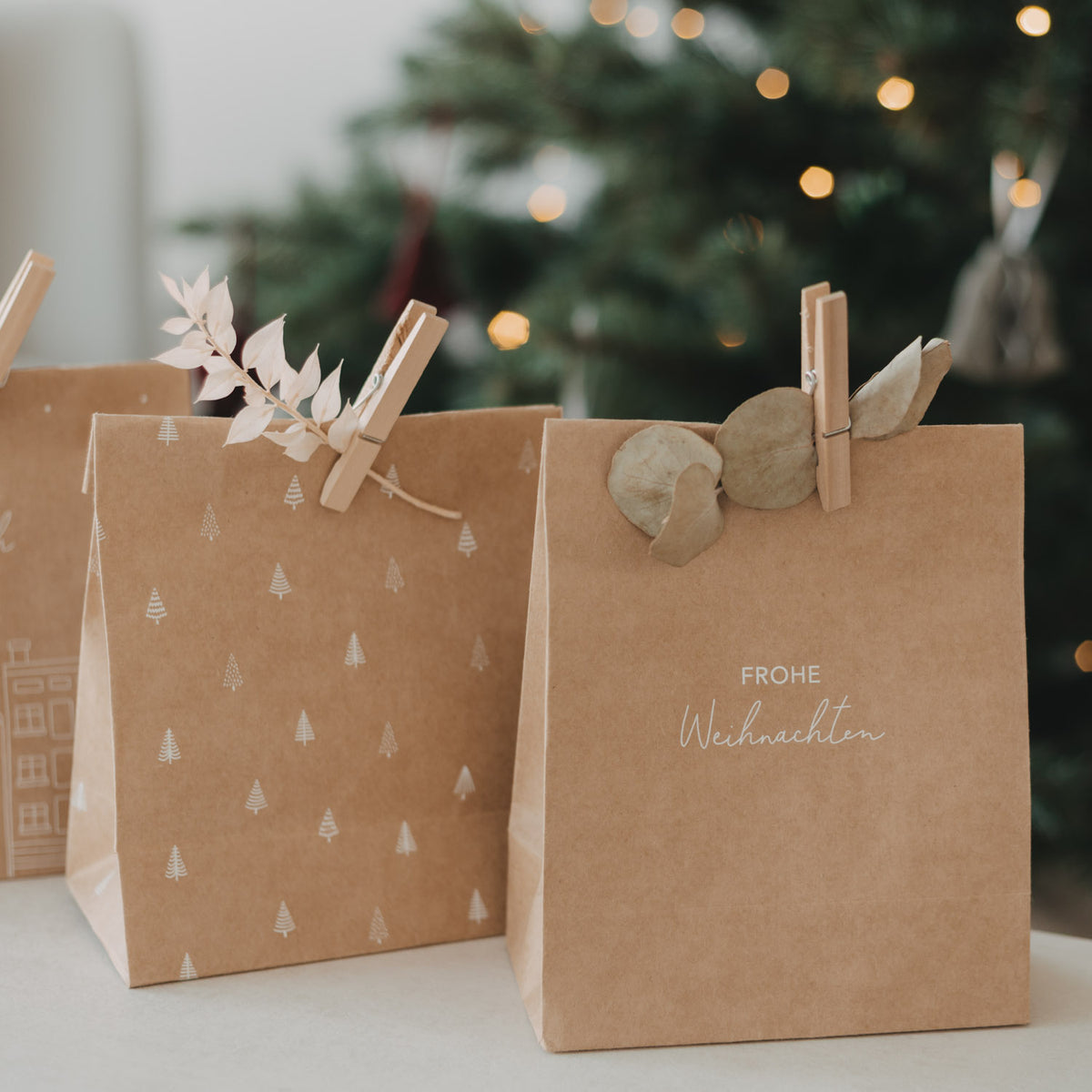 Braune Papiertüte mit Tannenmuster und Häusern neben Papiertüte „Frohe Weihnachten“